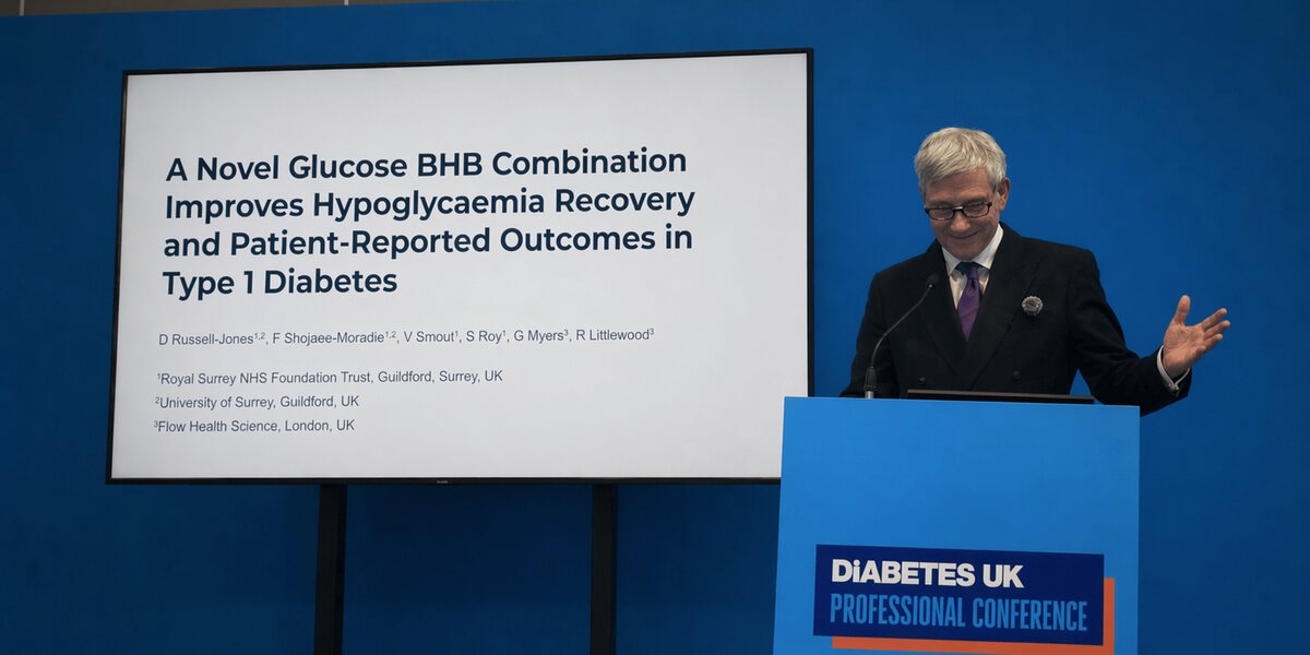 A speaker at the Diabetes UK Professional Conference podium beside a slide titled 'A Novel Glucose BHB Combination Improves Hypoglycaemia Recovery and Patient-Reported Outcomes in Type 1 Diabetes'